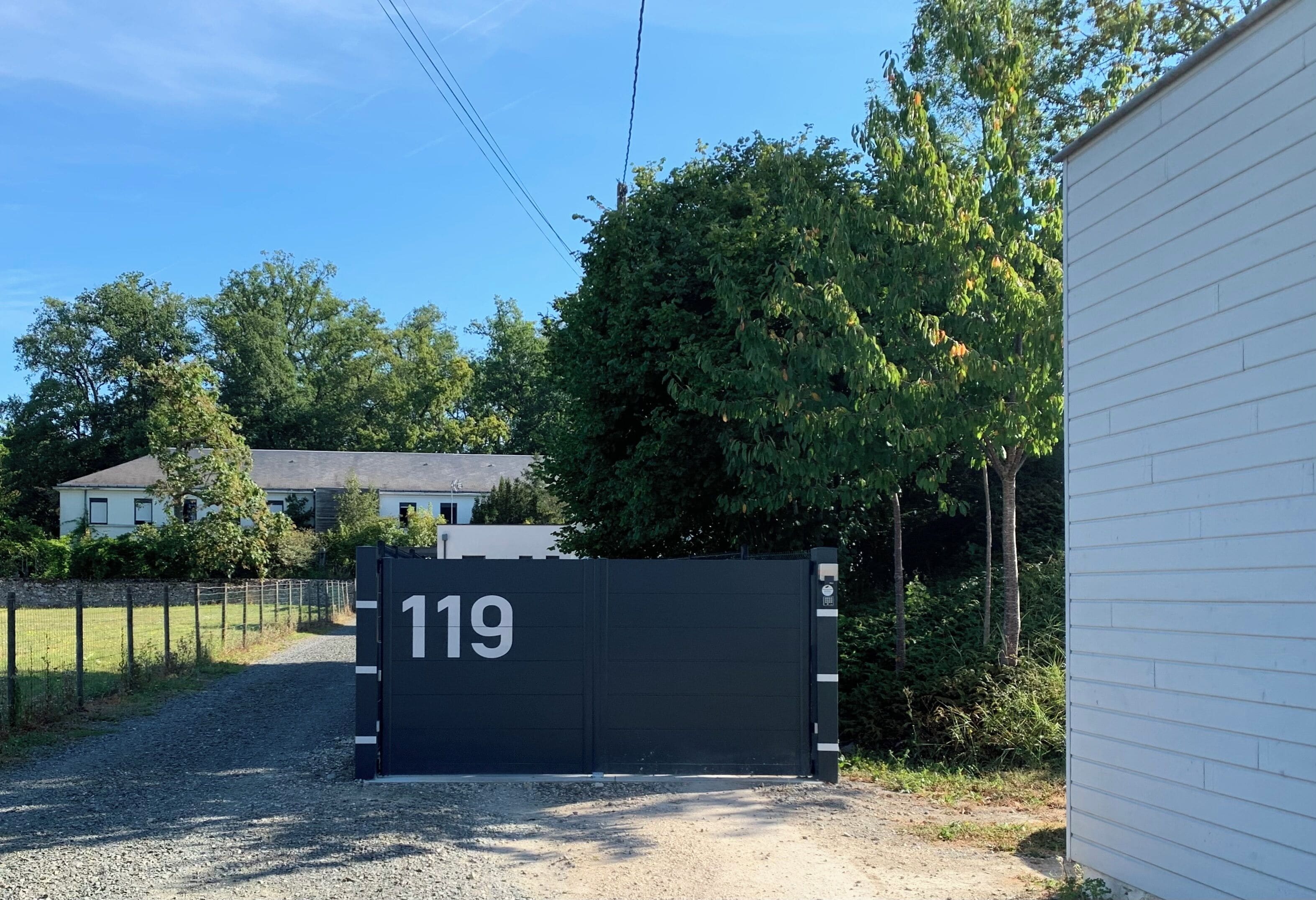 Een hekpoort met het nummer 119, een grindpad, een wit gebouw en bomen op de achtergrond.