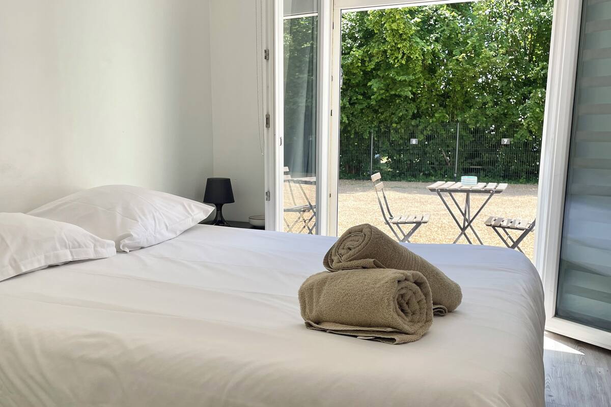 Una cama bien hecha con sábanas blancas y dos toallas enrolladas, una mesita con una lámpara, y vistas a un espacio exterior con sillas y una valla.
