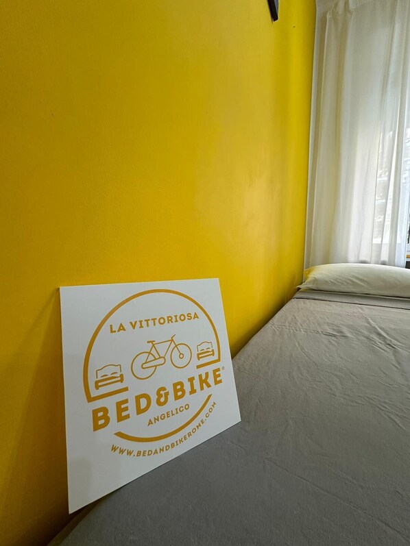 Una cama en una habitación con paredes amarillas y una cortina blanca. Un letrero en la pared dice "LA VITTORIOSA BED&BIKE ANGELICO".