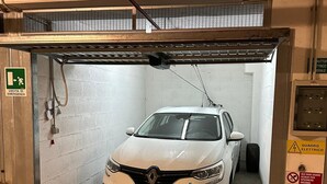 A white car parked inside a garage with a visible emergency exit sign and a warning sign about electrical hazards. - Bed&BikeRome ANGELICO  (Rome)