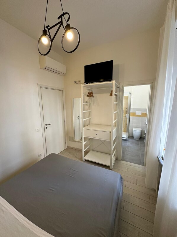 Quarto com cama, guarda-roupa, televisão e banheiro visível através de uma porta aberta.