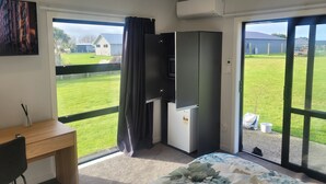 A bedroom with a bed, a desk, a chair, a wardrobe, and a large window with a view of a grassy area and a building. - A Gem In Ascot is situated in Invercargill. (Invercargill)