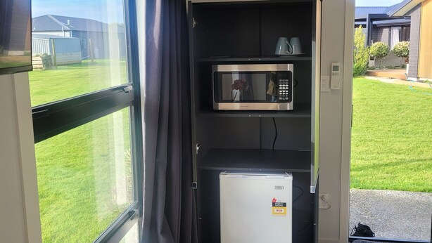 A compact kitchenette with a built-in microwave and refrigerator, a large window with a view of a grassy yard, and a ceiling-mounted air conditioning unit. - A Gem In Ascot is situated in Invercargill. (Invercargill)