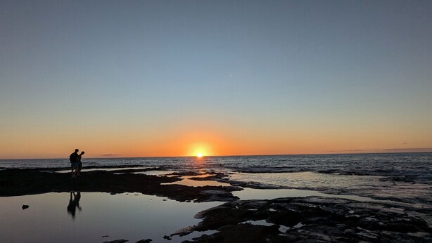 Atardecer sobre una playa rocosa con aguas tranquilas que reflejan el cielo.