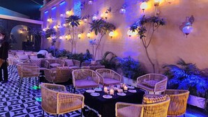 Un'area salotto all'aperto con sedie e tavoli in vimini, circondata da piante in vaso e illuminazione decorativa a parete.