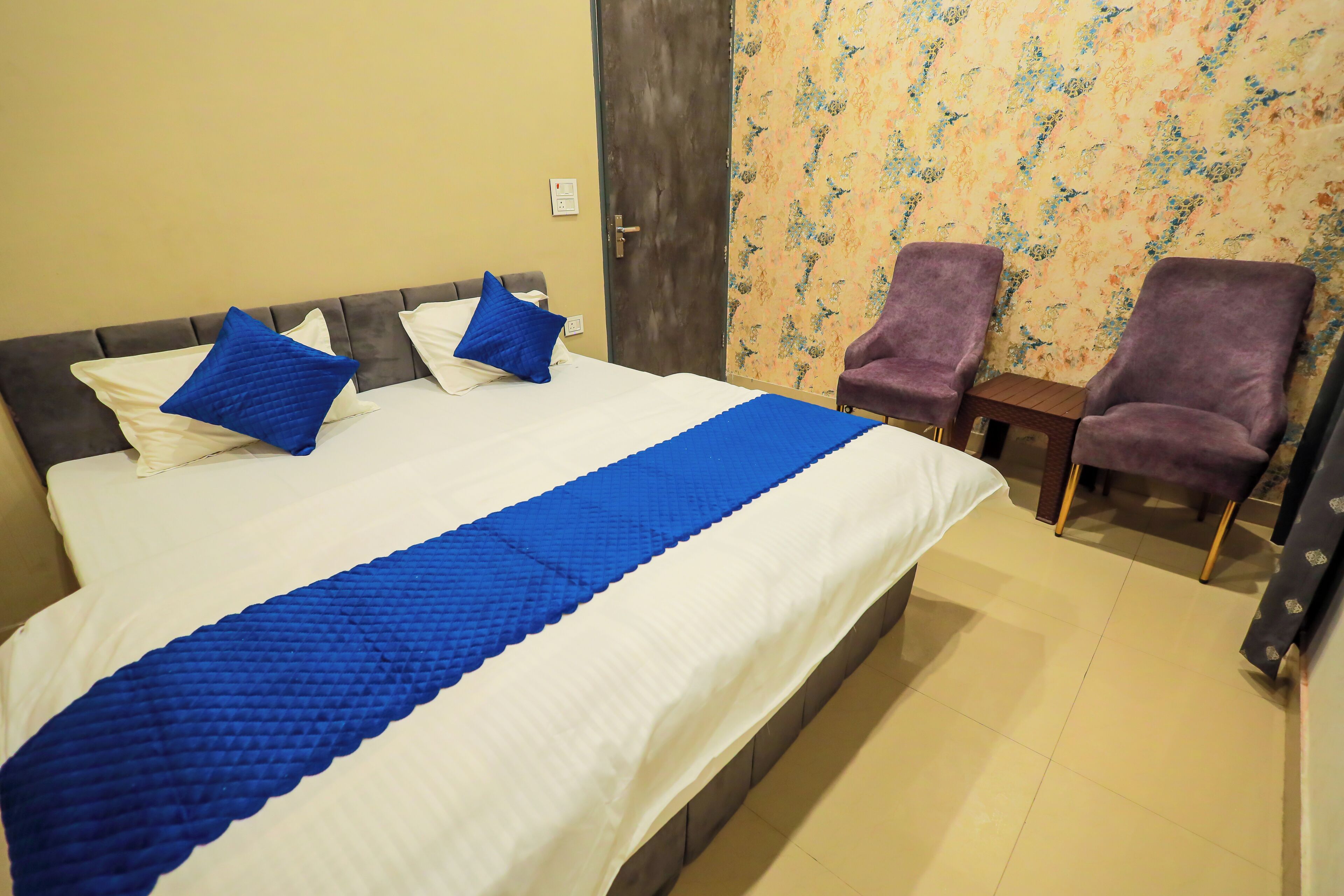 A hotel room with a large bed, two blue pillows, a blue bedspread, and two purple armchairs.