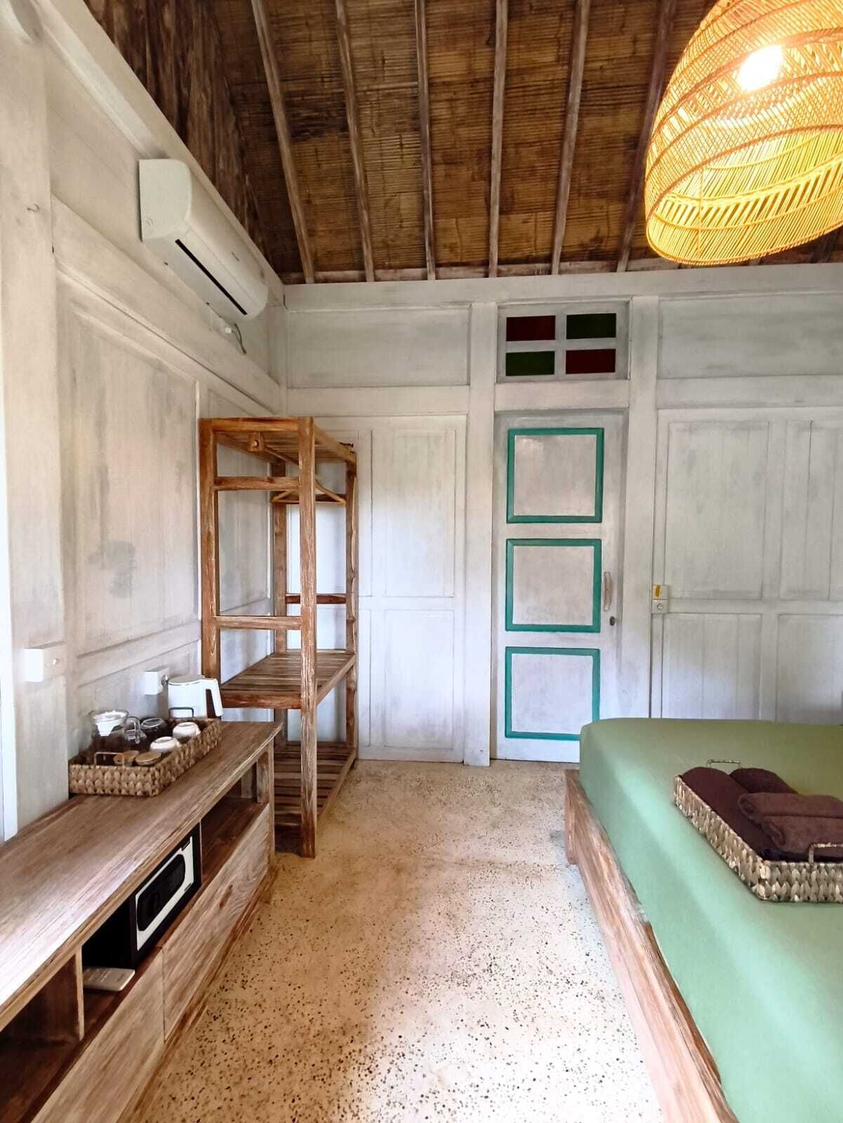 A room with a wooden ceiling, a bed with green bedding, a wooden bench, a shelf with items, and a door with green frames.