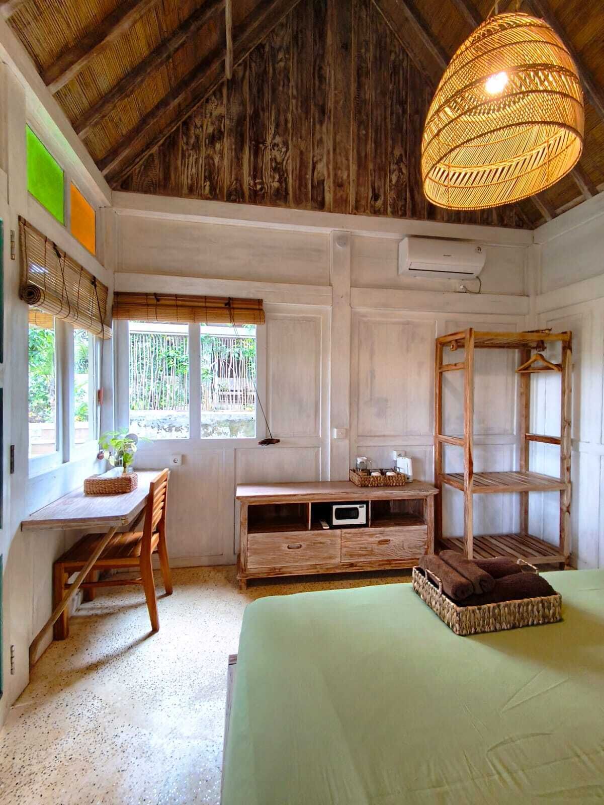 A rustic room with a wooden ceiling, a bed, a desk with a chair, a window with a view, and a hanging light fixture.