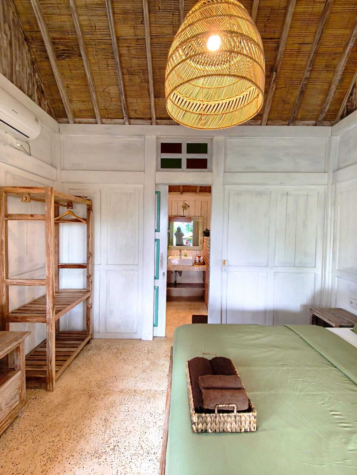 Una habitación con cama, lámpara colgante, repisa de madera y baño visible a través de una puerta abierta.