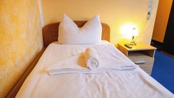 A neatly made bed with white linens and pillows, a rolled towel, a bedside table with a lamp, and a power outlet on the wall.