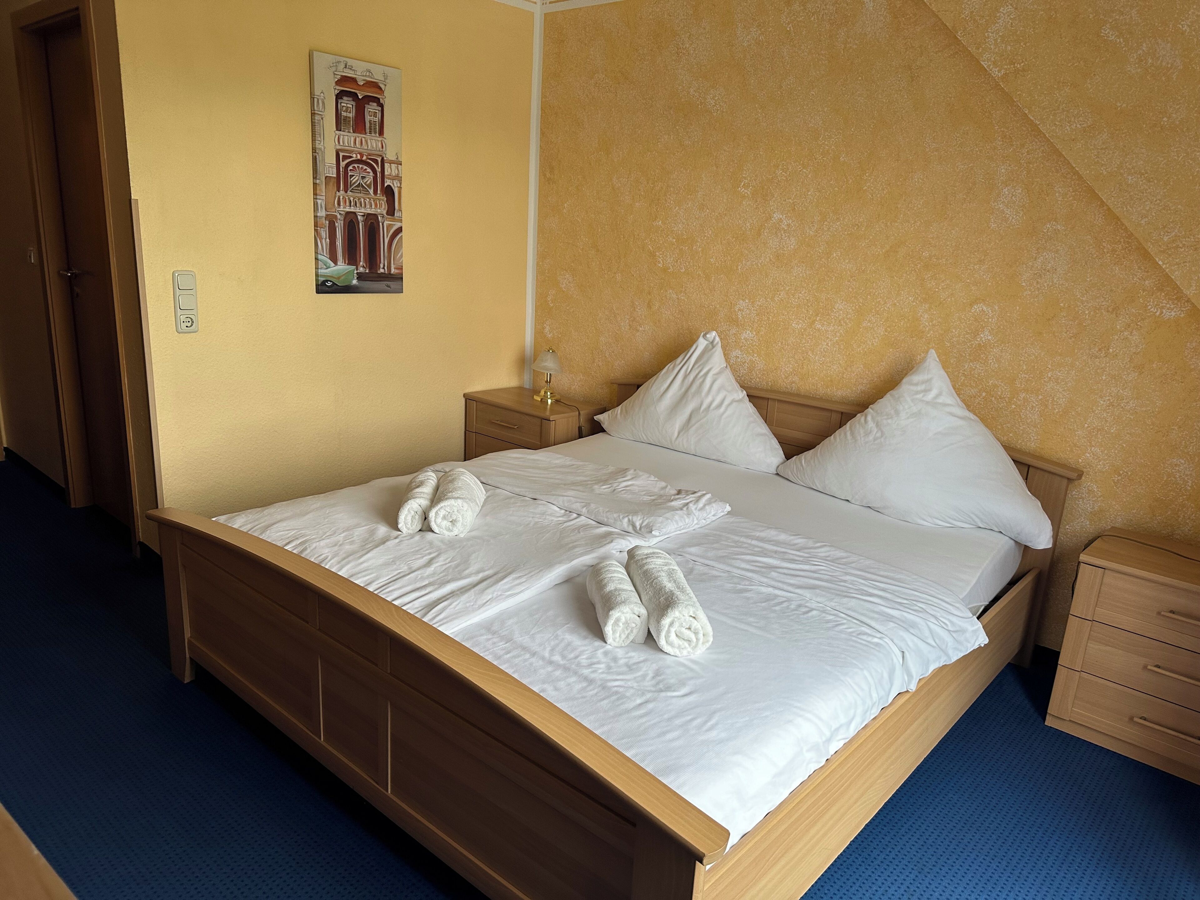 A neatly made bed with white linens and rolled towels, a wooden headboard, and a nightstand with a lamp.