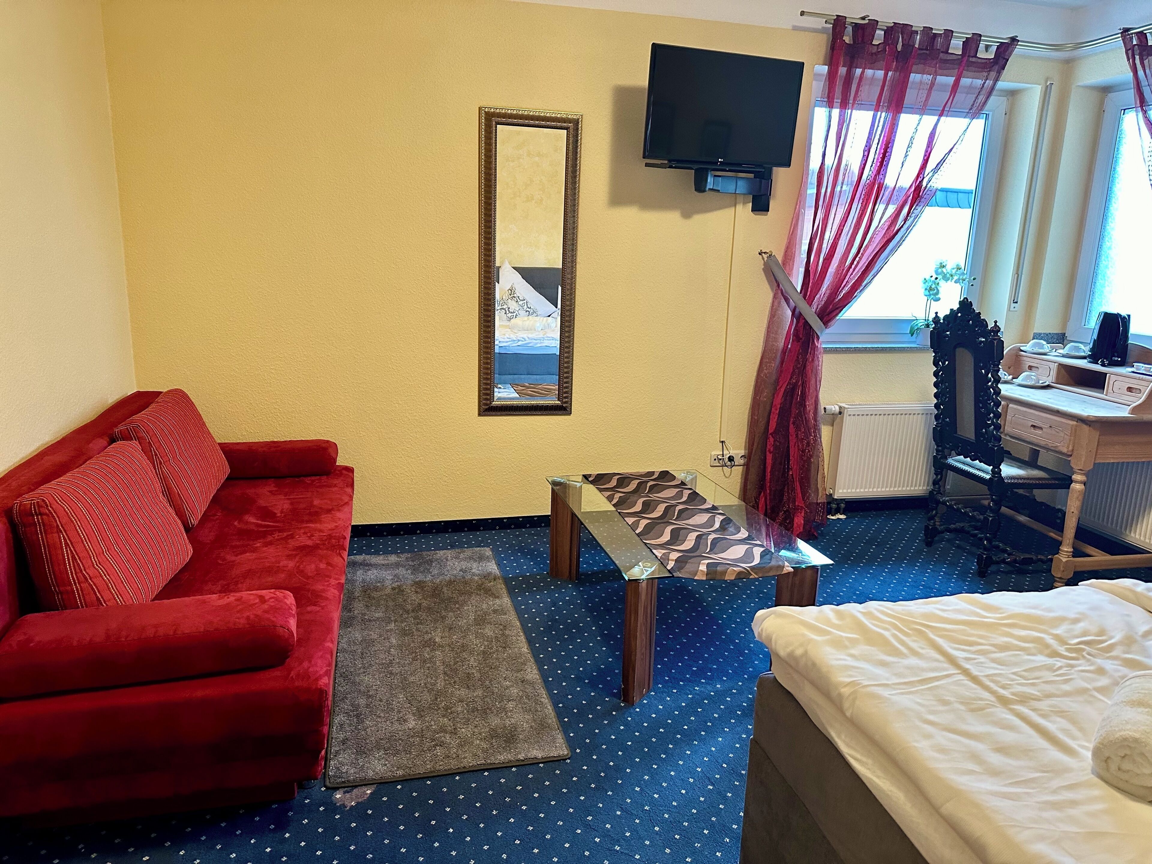 Una camera d'albergo con un divano rosso, un letto, una scrivania con un computer, una TV montata a parete e una finestra con tende.