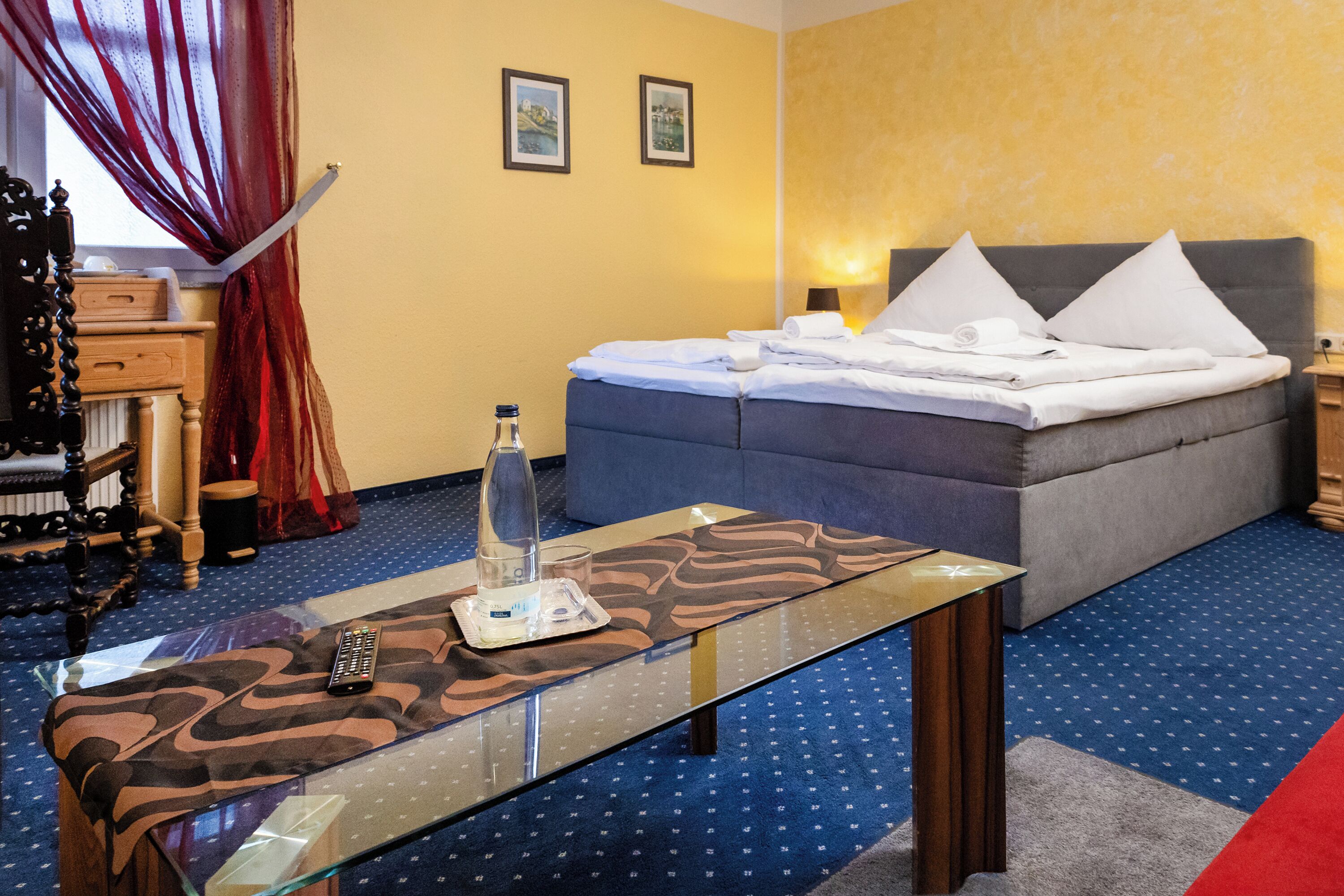 A hotel room with a bed, a bedside table with a remote control, a bottle, and a glass.
