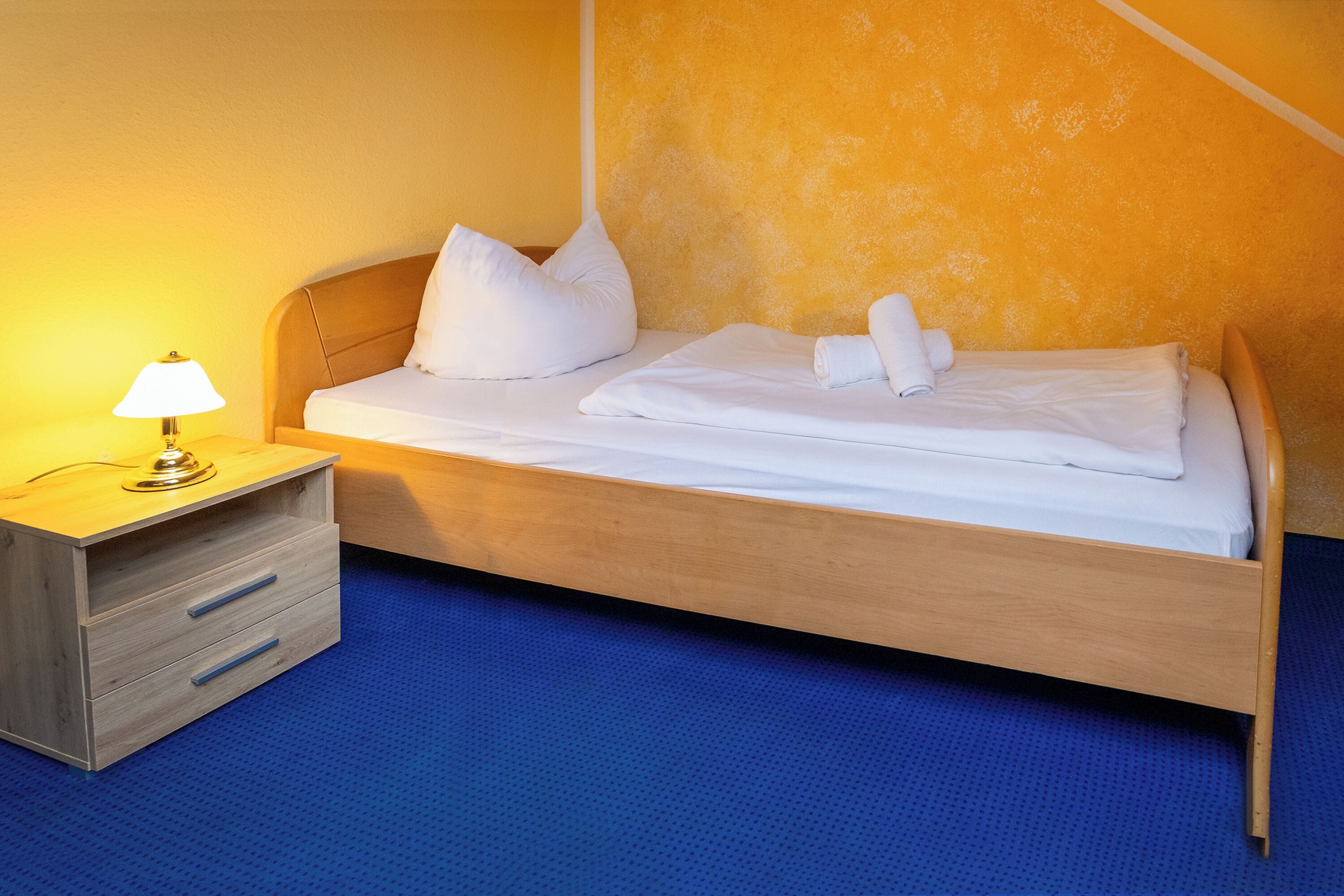Un lit bien fait, avec du linge de lit blanc et des oreillers, une table de chevet avec une lampe, et un sol recouvert d’une moquette bleue.