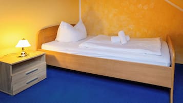 Una cama bien hecha con sábanas y almohadas blancas, una mesita de noche con lámpara y un suelo cubierto de alfombra azul.