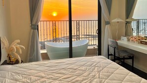 A hotel room with a large bed, a desk with a chair, a bathtub, and a balcony with a view of the sunset. - S Gen Residences Phu Quoc (Phu Quoc)