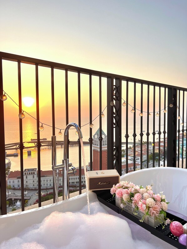 A balcony with a bathtub filled with bubbles, a flower arrangement, and a view of a cityscape with a bridge and buildings.