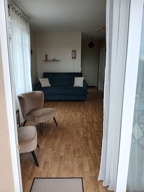 Un salon avec un canapé bleu, des fauteuils beiges, un parquet en bois et un mur blanc agrémenté d’une étagère décorative.