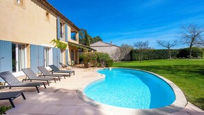 Una piscina con sillas de descanso, una casa con contraventanas azules y un césped cuidado.