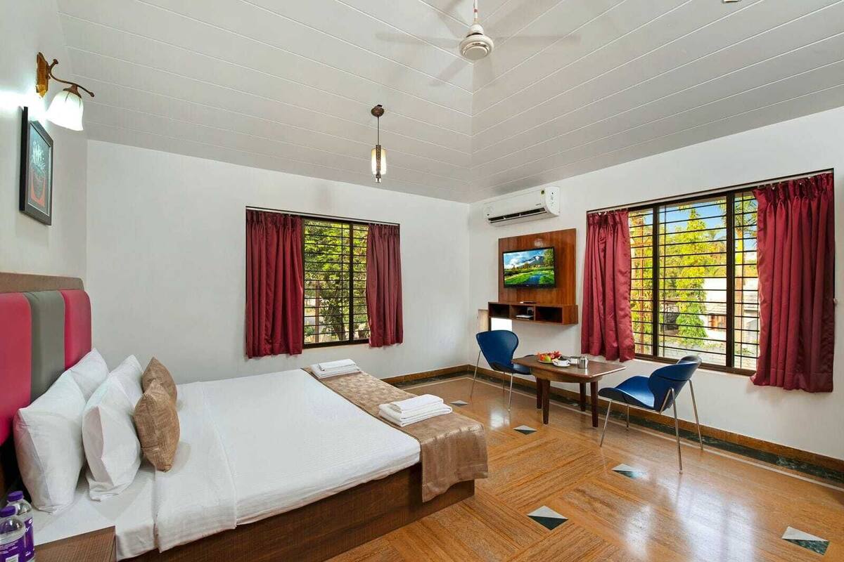 Une chambre d’hôtel avec un lit, un bureau, une chaise, une télévision et de grandes fenêtres avec des rideaux.