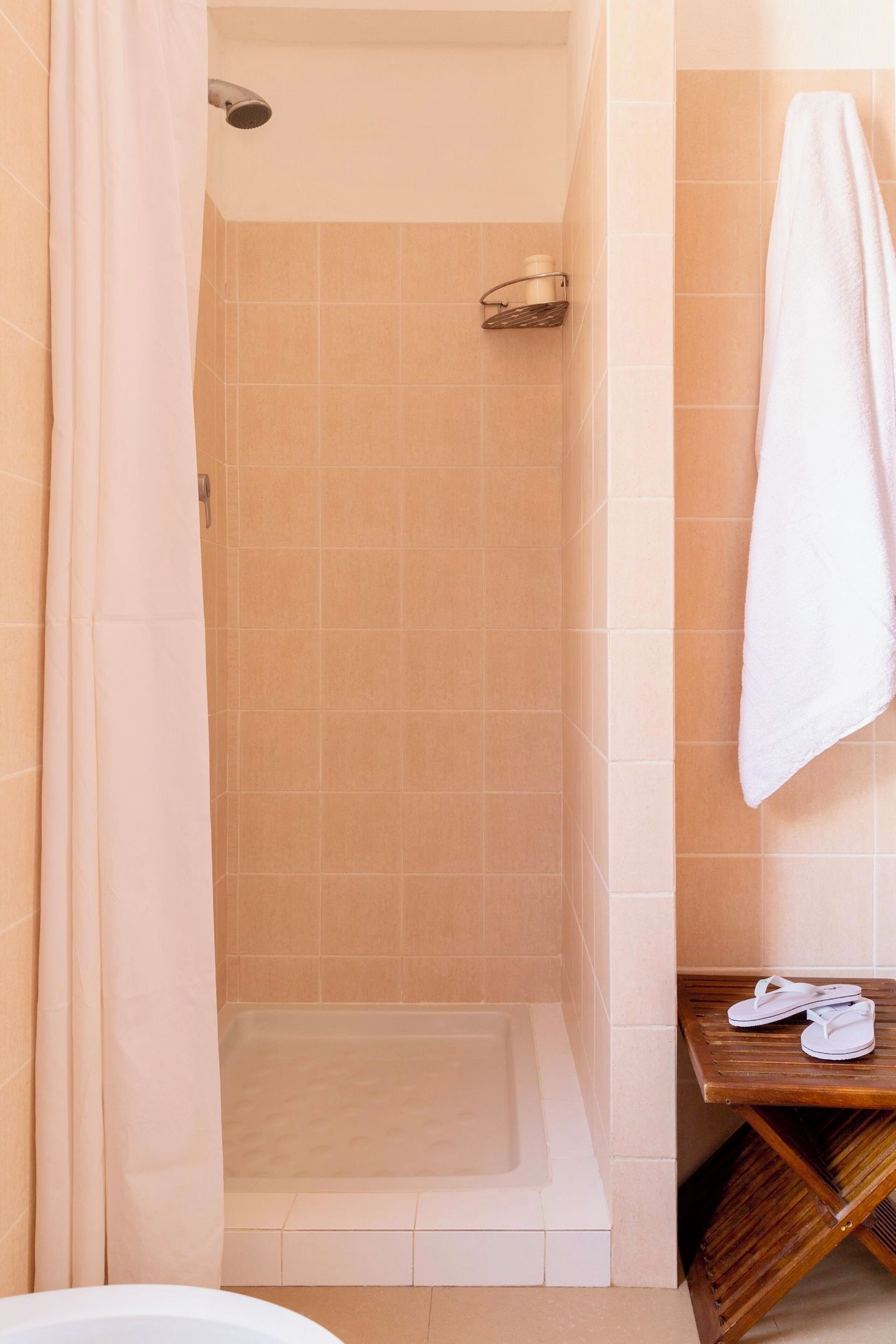 Une salle de bain avec une douche, une baignoire et une étagère en bois sur laquelle sont posés une serviette et des chaussons.