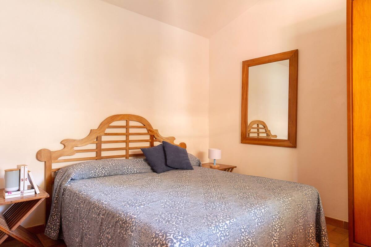 A bedroom with a wooden bed frame, a patterned bedspread, a bedside table with a lamp, and a mirror on the wall.