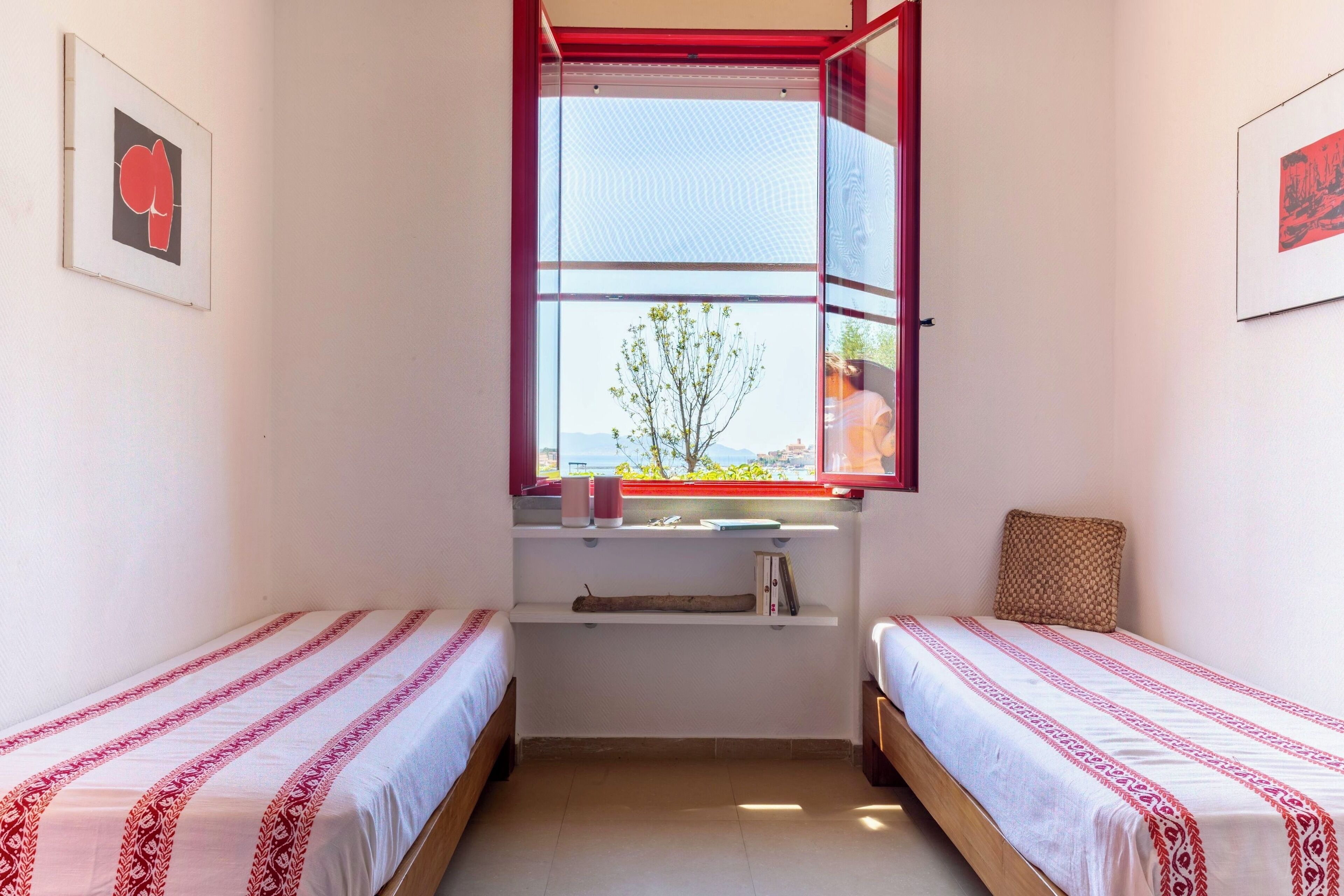 Una habitación pequeña con dos camas individuales, una ventana con vistas, y una estantería con libros y una vela.