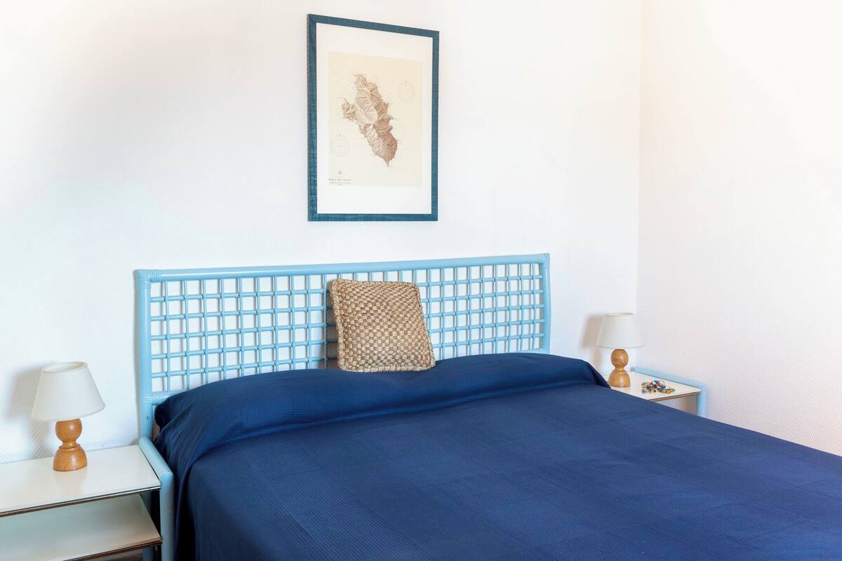 A bedroom with a blue headboard, a bed with a blue bedspread, a framed map on the wall, and two bedside tables with lamps.