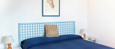 A bedroom with a blue headboard, a bed with a blue bedspread, a framed map on the wall, and two bedside tables with lamps.