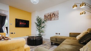 Una sala de estar con pared amarilla, un televisor de pantalla plana, una mesa de centro redonda y una planta grande.