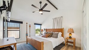 Un dormitorio con cama, ventilador de techo, mesita de noche con lámpara, ventana con cortinas y vistas al exterior.
