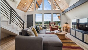 A spacious living area with a high ceiling, large windows, a sofa set, a coffee table, and a TV.
