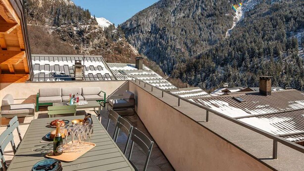 Una terraza con una mesa lista para comer, con vistas a un paisaje montañoso de picos nevados y bosques densos.