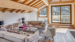 Un loft espacioso con techos altos y vigas de madera a la vista, que cuenta con una zona de estar acogedora con sofá, sillones y una mesa de centro. Amplias ventanas ofrecen vistas a un paisaje nevado.