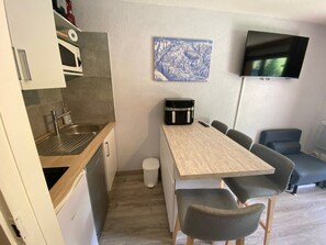 A compact kitchen with a sink, microwave, and a small trash bin. A wooden table with four chairs is positioned near a wall-mounted TV.