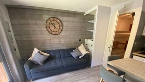 A compact living space with a sofa, a dining table with chairs, a clock on the wall, and a built-in kitchen area. - Cozy studio near ski area with parking (Châtel)