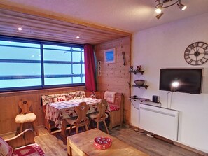A cozy cabin interior with a dining area, wooden walls, a large window, a clock, and a TV.