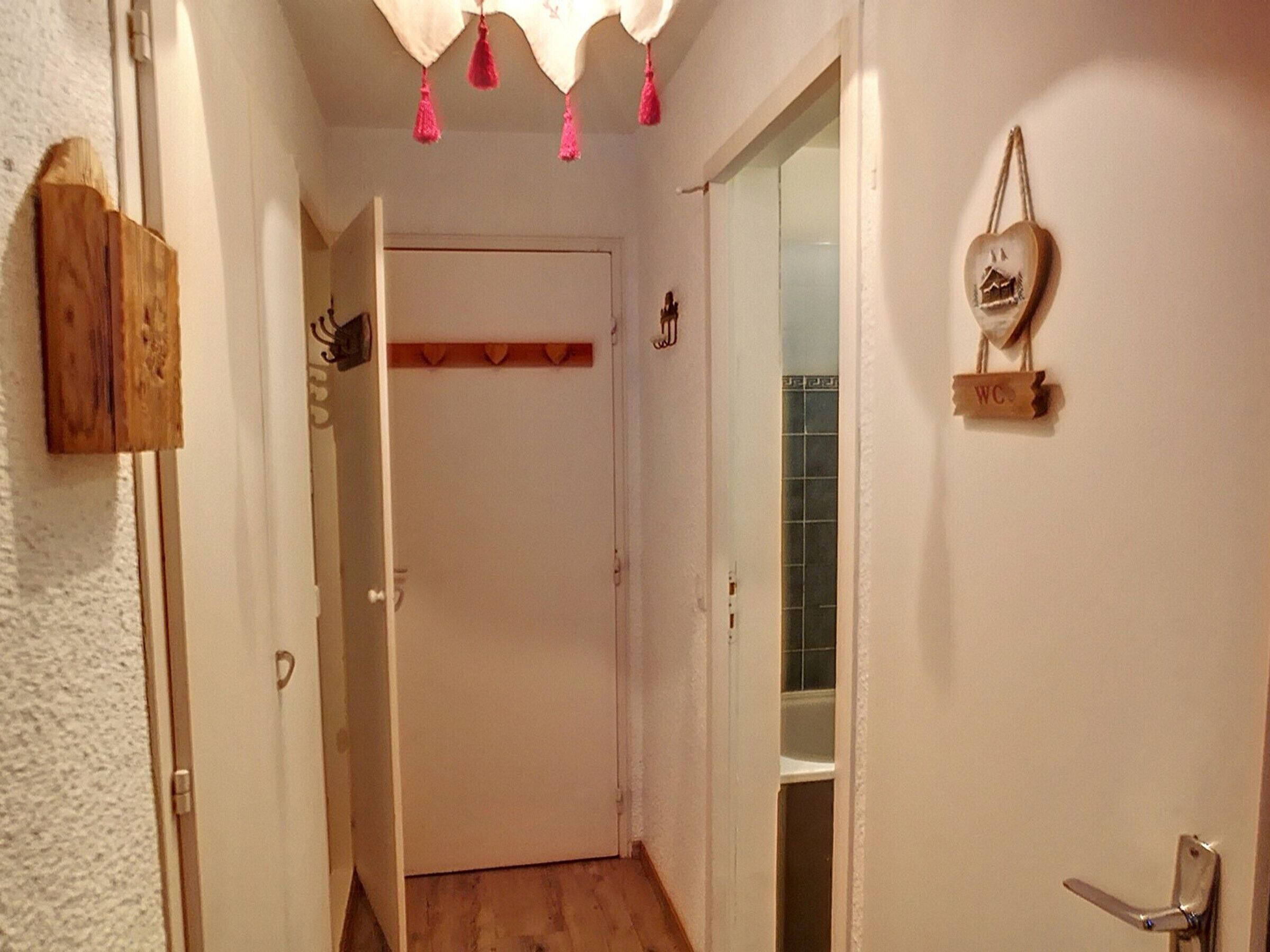 Een smalle gang met een houten deur aan het einde, een klein plankje met een decoratief object, en een bordje met een huissymbool.