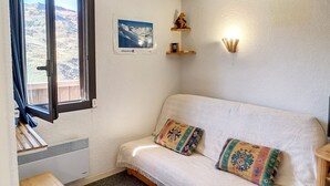 Um quarto aconchegante com um sofá branco, almofadas estampadas, uma janela com vista para montanhas, uma prateleira fixada na parede com livros e um arandela.