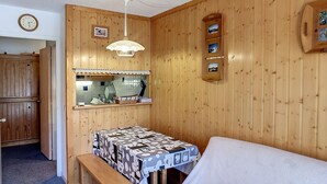 Um quarto acolhedor com paredes de madeira, uma cama, uma pequena mesa com toalha de mesa estampada, um banco, um relógio e um candeeiro fixado na parede.