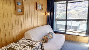 Quarto com paredes de madeira, cama com manta estampada, mesa pequena com toalha de mesa estampada, janela com vista para paisagem montanhosa e luminária no teto.