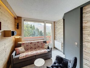 Una camera accogliente con una parete in legno, un divano con cuscini decorati, una poltrona nera e un'ampia finestra che si affaccia su alberi e montagne.