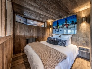 A bedroom with wooden walls, a large bed with white bedding, a knitted throw, and a ski-themed painting above the bed. - Charming apartment with included services in Courchevel (Courchevel)