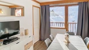 Une pièce avec une télévision, une table entourée de chaises, une fenêtre avec des rideaux et une vue sur un paysage enneigé.