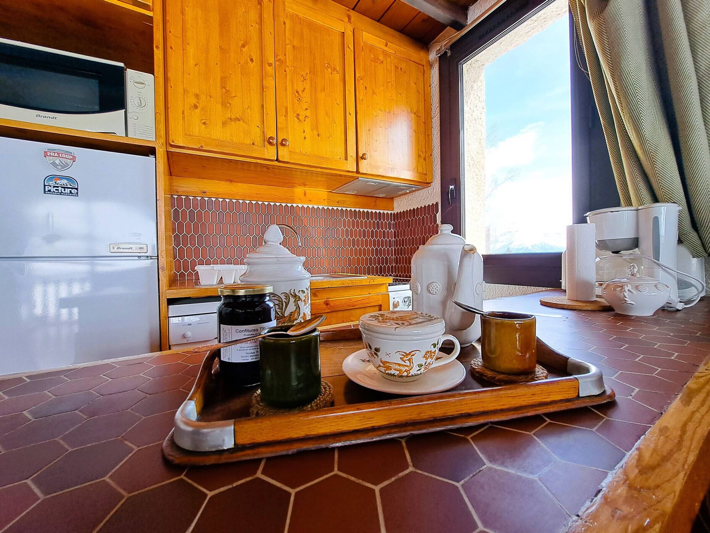 A kitchen with wooden cabinets, a microwave, a refrigerator, a coffee maker, and a tray with a teapot, cups, and a jar of jam.