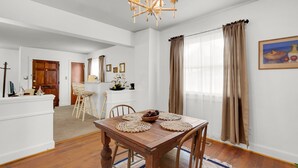 Ein Essbereich mit Holztisch, Stühlen und einer Chandelüster. Im Hintergrund ist eine Küche mit Holzschränken und einer weißen Arbeitsplatte zu sehen.
