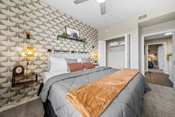 Un dormitorio con papel tapiz estampado, una cama con manta gris y marrón, lámparas de noche y un reloj en un estante.