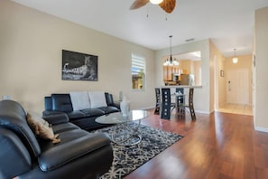 A living room with a black leather sofa, a glass coffee table, and a patterned rug. There is a dining area with a table and chairs, and a kitchen in the background.