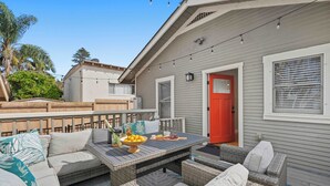 Un patio al aire libre con mobiliario de mimbre, una mesa de comedor con frutas y un cuenco, y una puerta roja.
