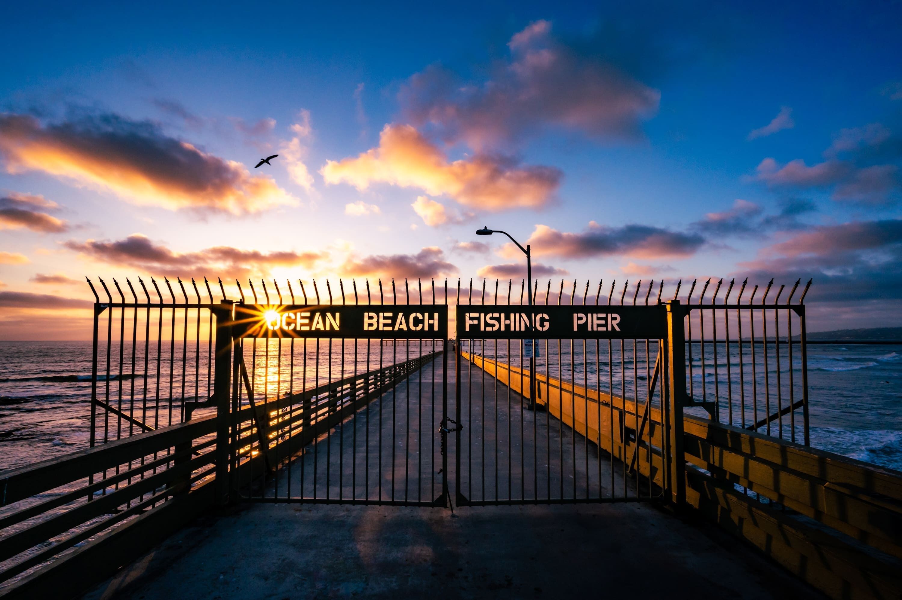 A pier with a gate labeled 'Ocean Beach Fishing Pier' and a sunset in the background.